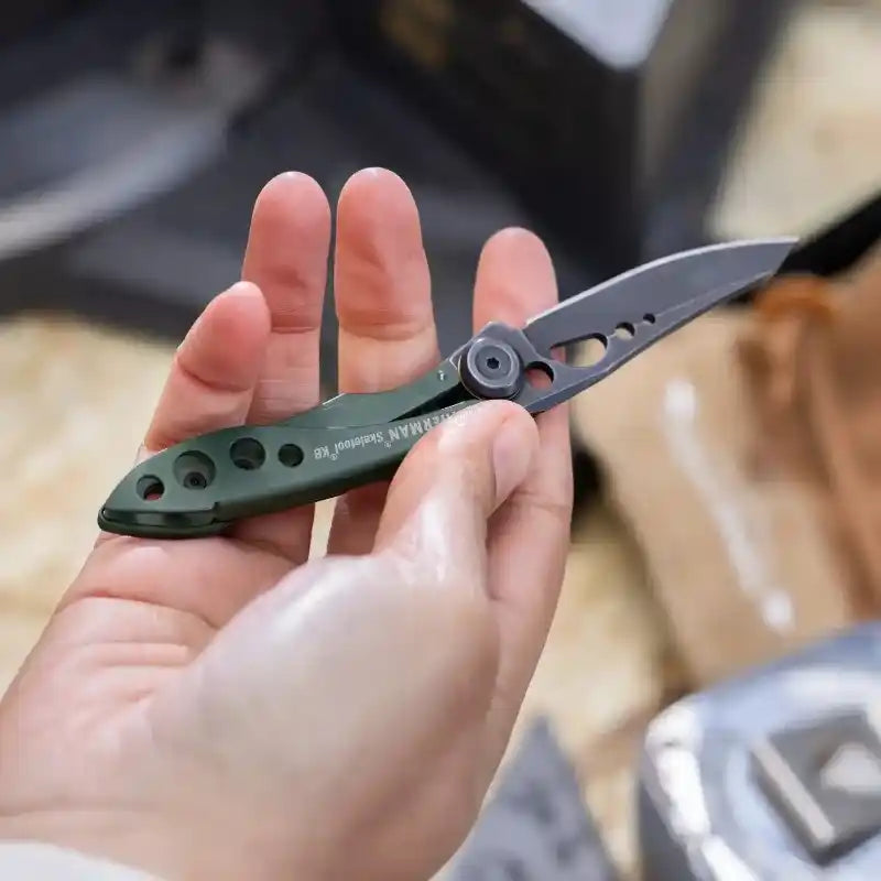 Una mano humana sostiene una navaja Leatherman Skeletool KB abierta, de color verde oliva y negro